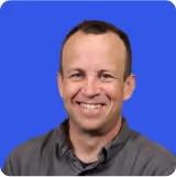 Smiling man with short brown hair wearing a dark gray collared shirt against a blue background.